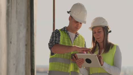 Ingenieros-Diseñadores-Un-Hombre-Y-Una-Mujer-Parados-En-El-Techo-De-Un-Edificio-En-Construcción-Y-Discuten-El-Plan-Y-El-Progreso-De-La-Construcción-Usando-Una-Tableta-Y-Un-Teléfono-Móvil.-Los-Constructores-Modernos-Discuten-La-Infraestructura-Del-Edificio-Y-Sus-Alrededores.